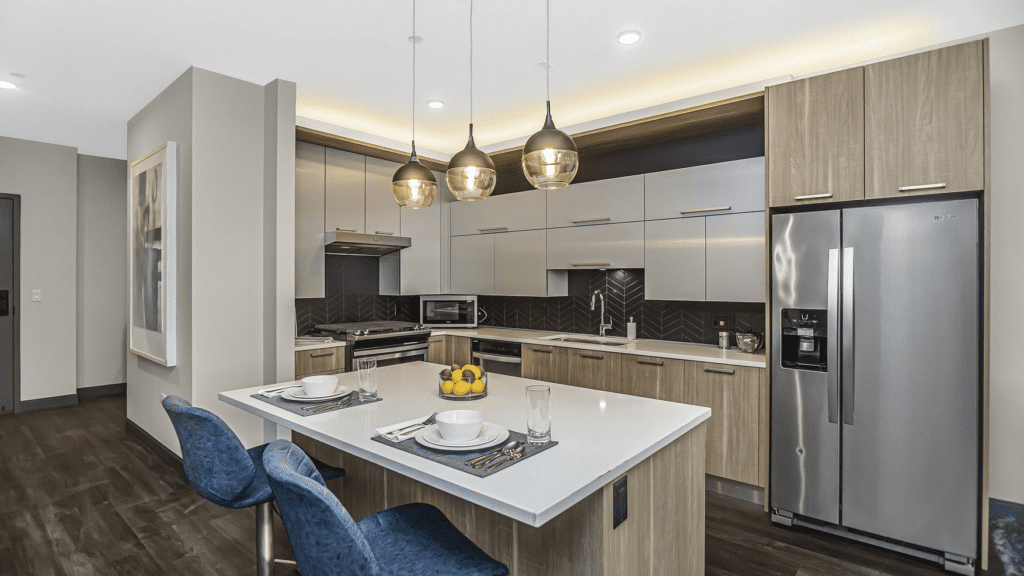 Modern kitchen with wooden cabinets, stainless steel appliances, and a white island counter. Three pendant lights hang above, with two blue chairs at the island. A bowl of fruit is on the counter.