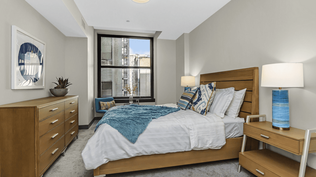 A modern bedroom with a wooden bed, blue blanket, patterned pillows, bedside tables with lamps, a wooden dresser, a potted plant, and a window with a view outside.
