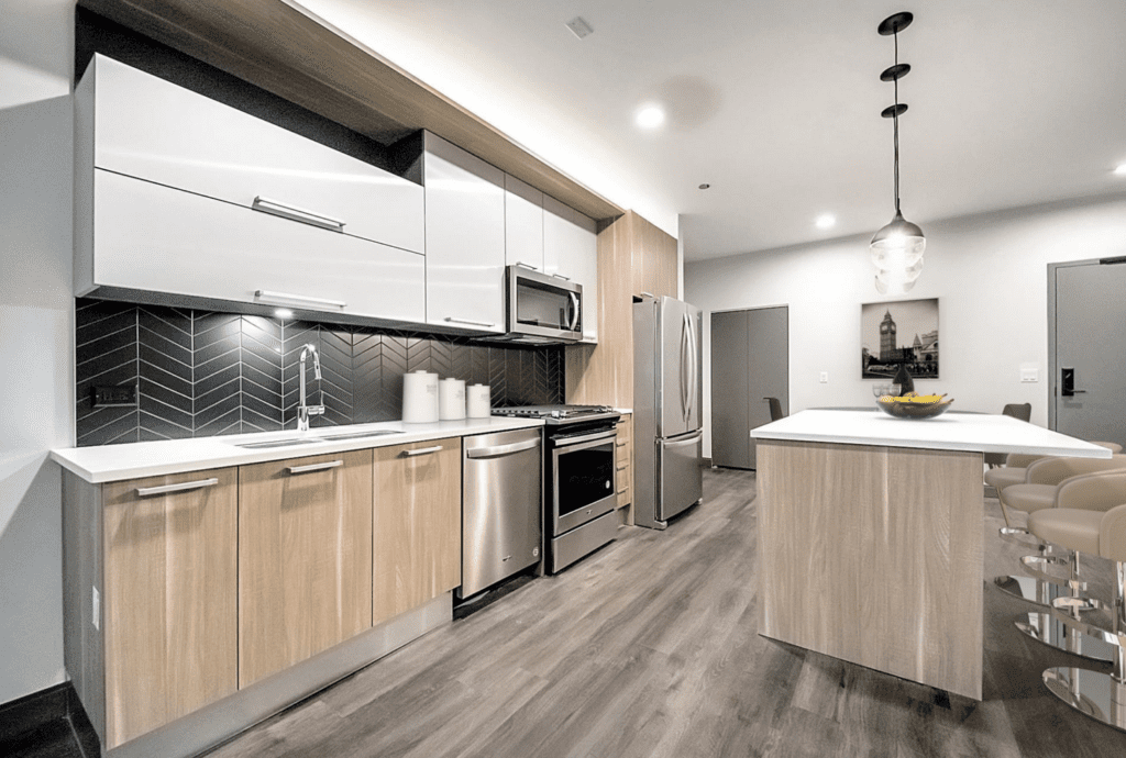 Modern kitchen with wooden cabinets, stainless steel appliances, black herringbone backsplash, and a white island with stools. A decorative photo and fruit bowl are on the counter.