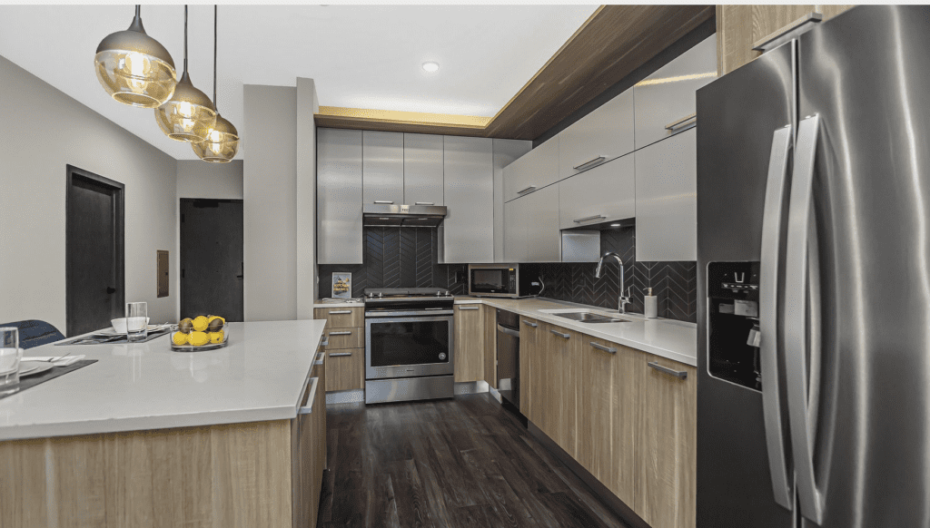 Modern kitchen with stainless steel appliances, wooden cabinets, and a dark herringbone backsplash. Hanging pendant lights illuminate the white countertops and fruit bowl.