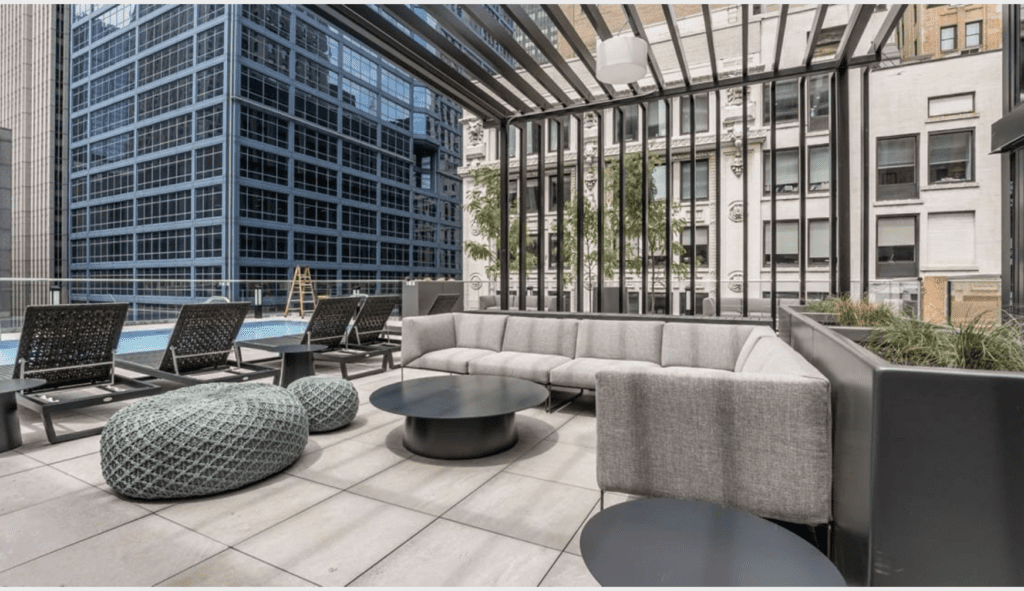 Rooftop lounge with gray sofas, round tables, woven ottomans, and loungers by a pool, surrounded by tall urban buildings.