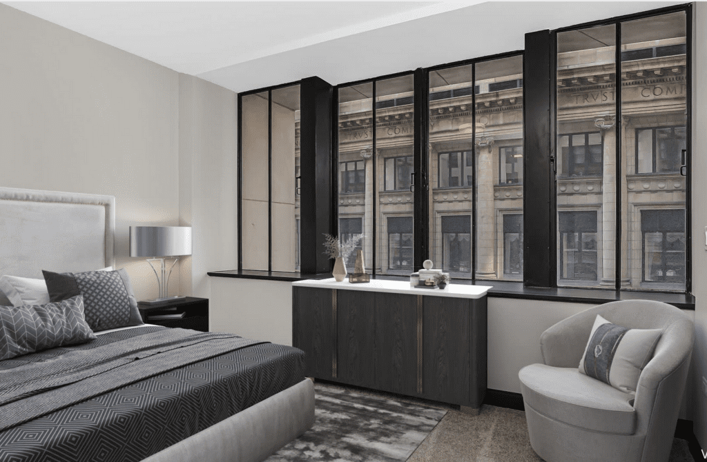 Modern bedroom with a bed, cushion chair, and a console table by tall windows overlooking an urban building. Neutral tones and geometric patterns accentuate the decor.