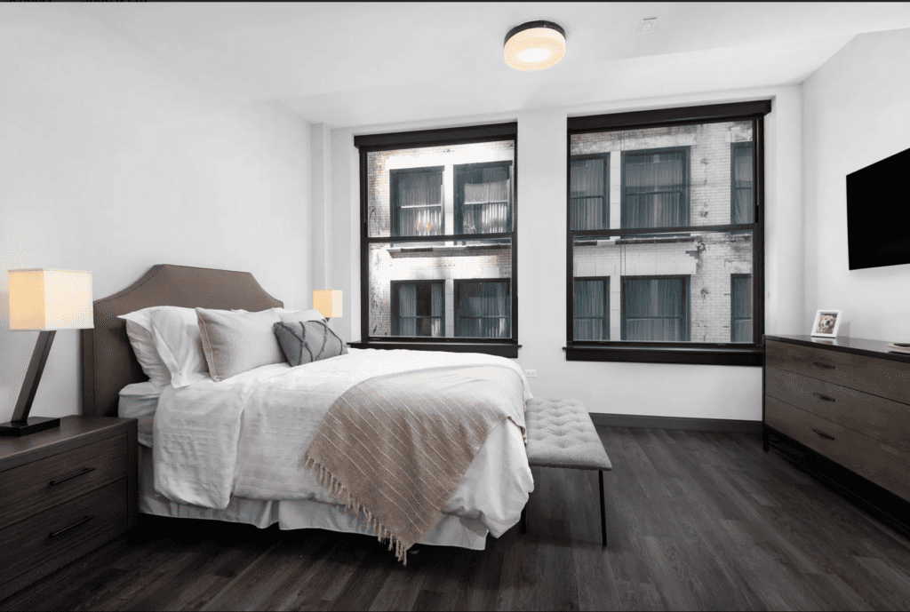 A modern bedroom with a neatly made bed, two nightstands with lamps, a bench at the foot of the bed, a dresser with a TV on the wall, and large windows showing a neighboring building.