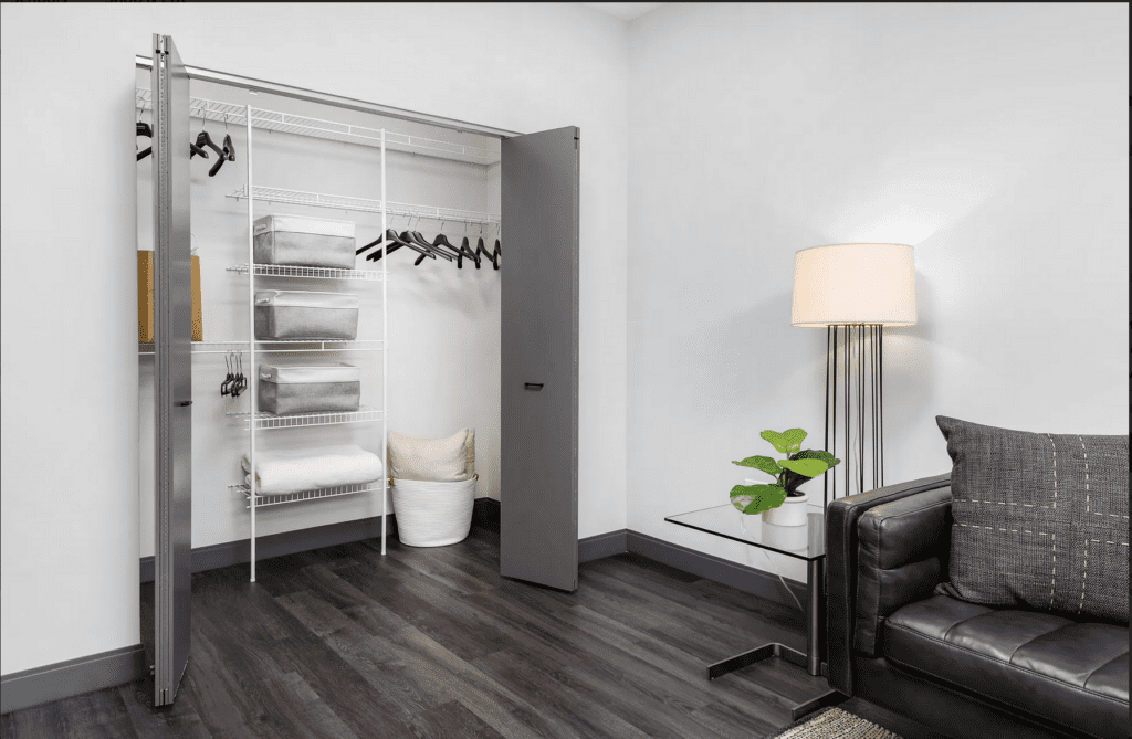 A modern interior with a partially open closet containing shelves, hangers, and linens. A black sofa, glass side table, lamp, and potted plant are in the foreground.