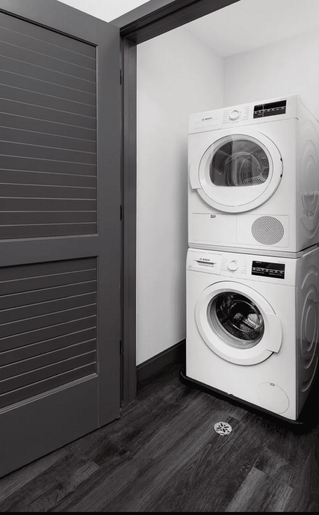 Stacked white washer and dryer in a small laundry nook with dark wood flooring and a gray door.