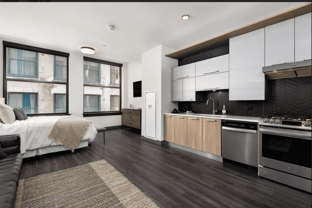 Modern studio apartment with a bed, sofa, and kitchen area. Dark wood flooring, white and wood cabinetry, and black backsplash. Large windows provide natural light. Neutral decor tones.