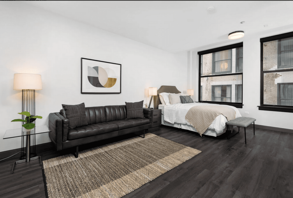 A modern studio apartment with a black sofa, glass coffee table, and bed. A woven rug is on dark wood floors. Walls are white with abstract art and large windows. A floor lamp stands nearby.