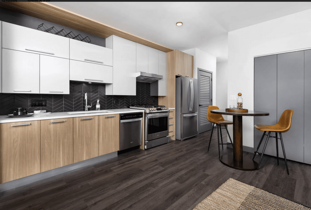 Modern kitchen with wooden and white cabinets, stainless steel appliances, dark herringbone backsplash, round table, and tall mustard-yellow chairs on a dark wood floor.