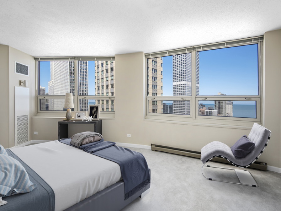 Modern bedroom with a large bed, a contemporary lounge chair, and a desk with a lamp. Tall windows offer a city view with high-rise buildings and a glimpse of the water in the background.