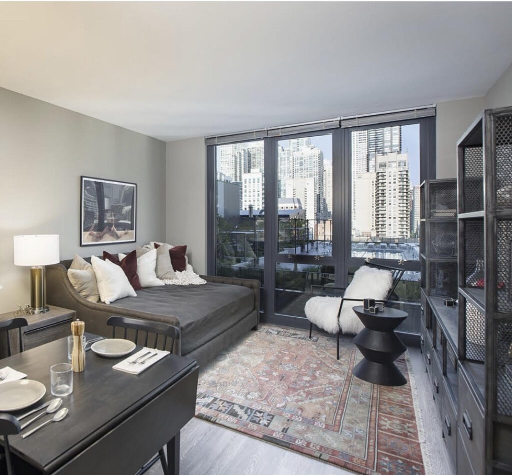 Modern living room with a bed and dining area. Large windows with city views, dark furniture, a patterned rug, and a framed photo on the wall.