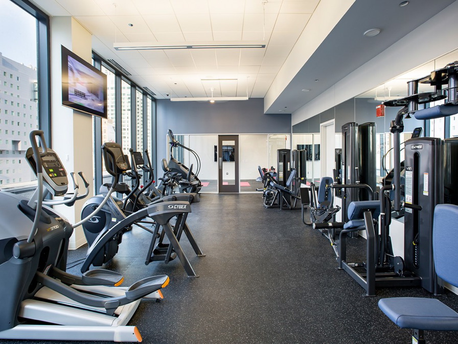 A modern gym with various exercise equipment, including treadmills, ellipticals, and weight machines. Large windows provide natural light.