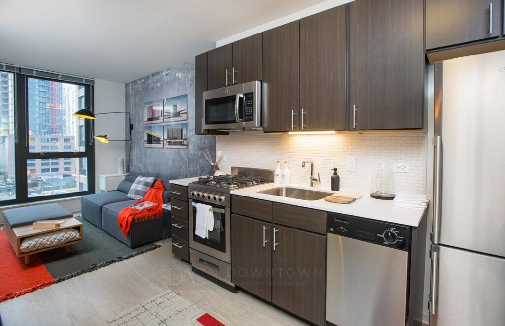 Modern kitchen and living area with dark wood cabinets, stainless steel appliances, and a small sofa by a large window. Urban view outside.