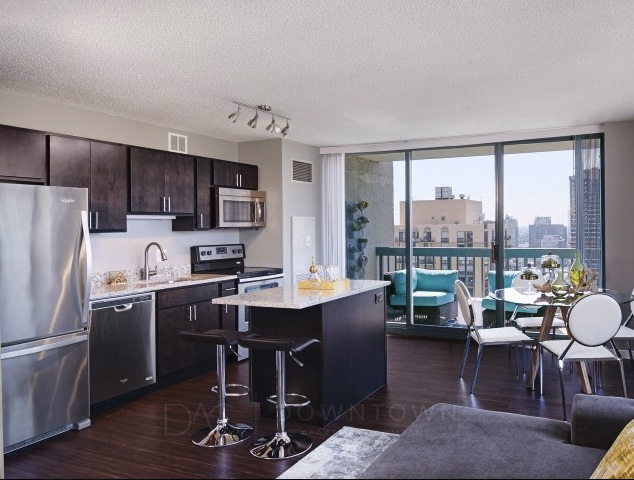 Modern apartment interior with a kitchen featuring dark wood cabinets, stainless steel appliances, a central island, and an adjacent living area with a dining table and city view through large windows.