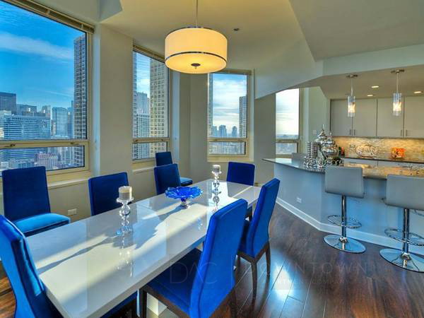 Modern dining area with a white table, blue chairs, and a view of city skyscrapers through large windows. Kitchen with bar stools and overhead lighting visible in the background.