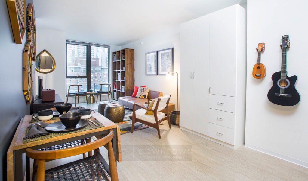 Modern apartment interior with a dining area, living space with a sofa, and guitars on the wall. Large window and shelves with books in the background. Light wood flooring throughout.