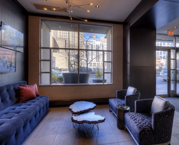 Modern hotel lobby with a blue tufted sofa, two dark patterned chairs, and an abstract coffee table. Large window and glass door reveal urban buildings outside. Neutral color palette.