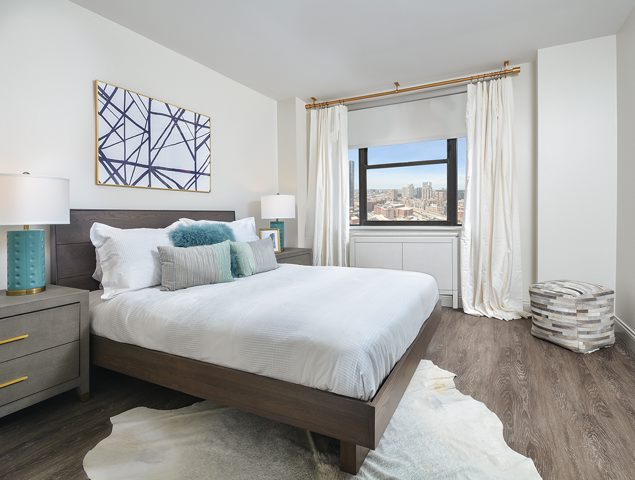 Modern bedroom with a large bed, abstract art, and two side lamps. A window with a city view is framed by white curtains. A small ottoman sits in the corner. Wood flooring complements the decor.