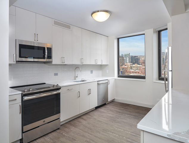 Modern kitchen with white cabinets, stainless steel appliances, and a large window overlooking a cityscape. Wood flooring and marble countertops add a sleek finish.