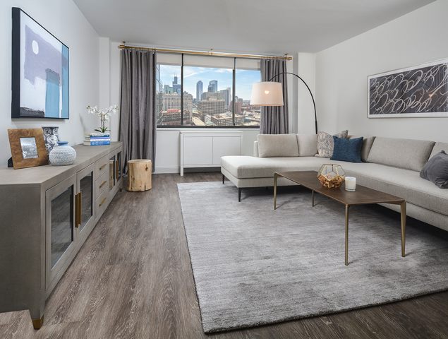 Modern living room with a gray sectional sofa, a wooden coffee table, a large area rug, and cityscape view through a window. Decor includes framed art and vases.