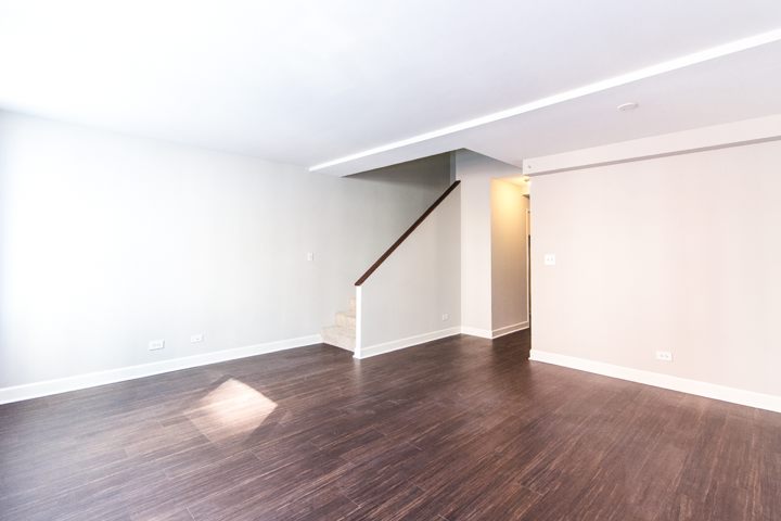 Empty room with dark wood flooring, light gray walls, and a staircase leading upstairs. Natural light shines from a window on the left.