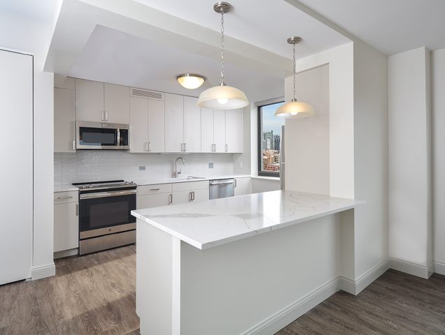 Modern kitchen with white cabinets, stainless steel appliances, marble countertop, pendant lights, and wood flooring, overlooking a cityscape through a large window.