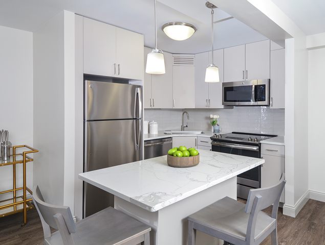 Modern kitchen with white cabinets, stainless steel appliances, and a marble island. Two stools are at the island, and a bowl of green apples is centered on it. Pendant lights hang above.