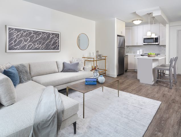 Modern living room and kitchen with a white sectional sofa, gray rug, and a sleek coffee table. Kitchen features white cabinetry, stainless steel appliances, and a small island with barstools.