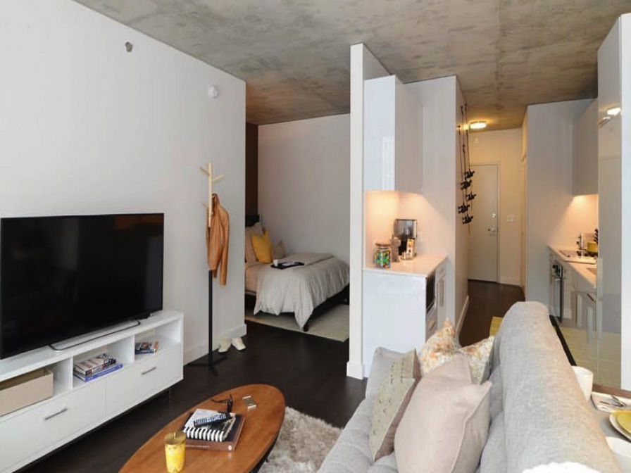 Modern studio apartment with a living area featuring a sofa, TV, and coffee table. The bedroom area shows a bed and a coat rack. The kitchenette and entrance are visible in the background.