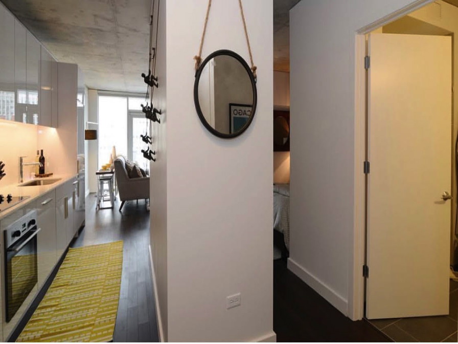 Modern apartment interior with a kitchen on the left featuring white cabinetry and wall-mounted wine rack, a hallway with a round mirror, and an open door leading to another room on the right.