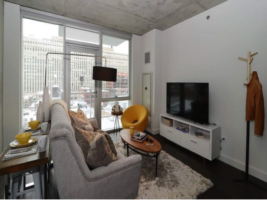 Modern living room with a grey sofa, yellow chair, TV on a white stand, and a window showing city buildings. Dining table with two place settings is partially visible.