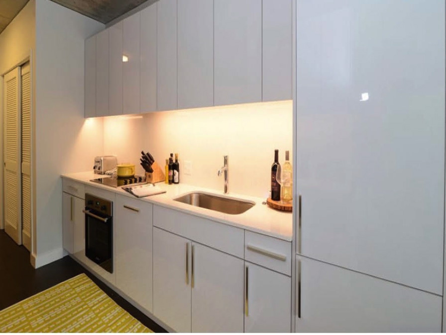 Modern kitchen with white cabinets, stainless steel sink, and an electric stove. Countertop features a knife block, toaster, and bottles. Bright under-cabinet lighting.