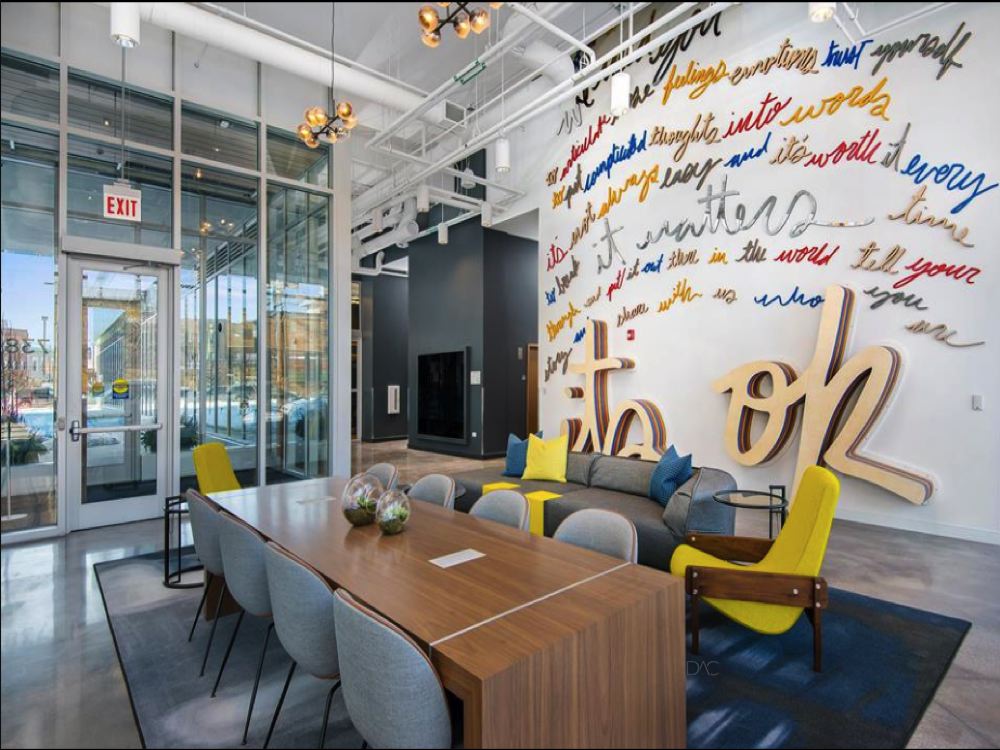 Modern office space with a wooden table, gray chairs, yellow armchairs, and a wall with colorful, cursive text art. Glass doors and windows let in natural light.