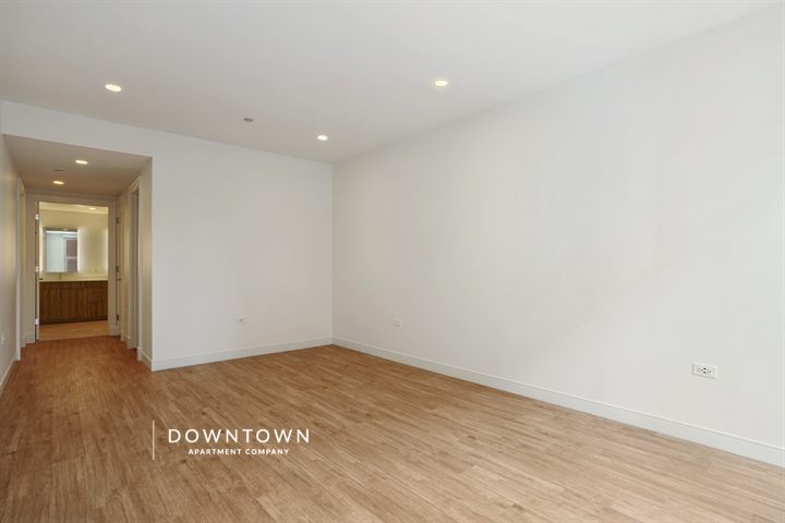 Empty room with light wood flooring, white walls, and recessed lighting. Hallway with a doorway leads to another area. Subtle branding text in the corner.