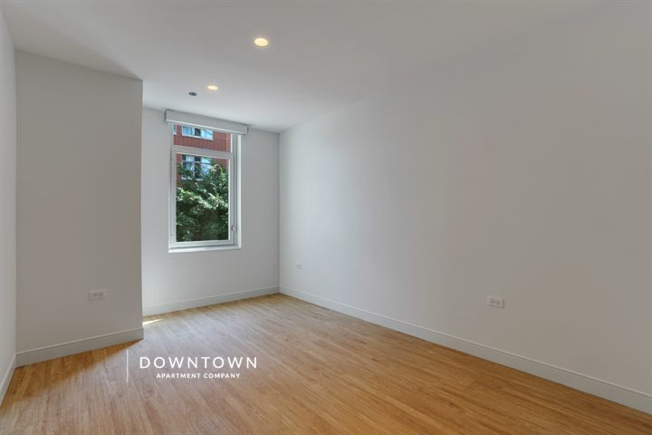 Empty room with light wood flooring, white walls, and a large window showing a partial view of a red brick building and greenery. Ceiling lights are on, and a logo reads "DOWNTOWN APARTMENT COMPANY.