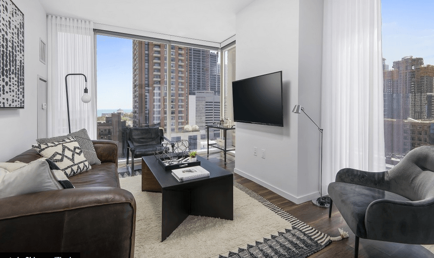 Modern living room with a brown leather sofa, black chairs, and a wall-mounted TV. Large windows offer a cityscape view. White curtains and a geometric rug complete the decor.