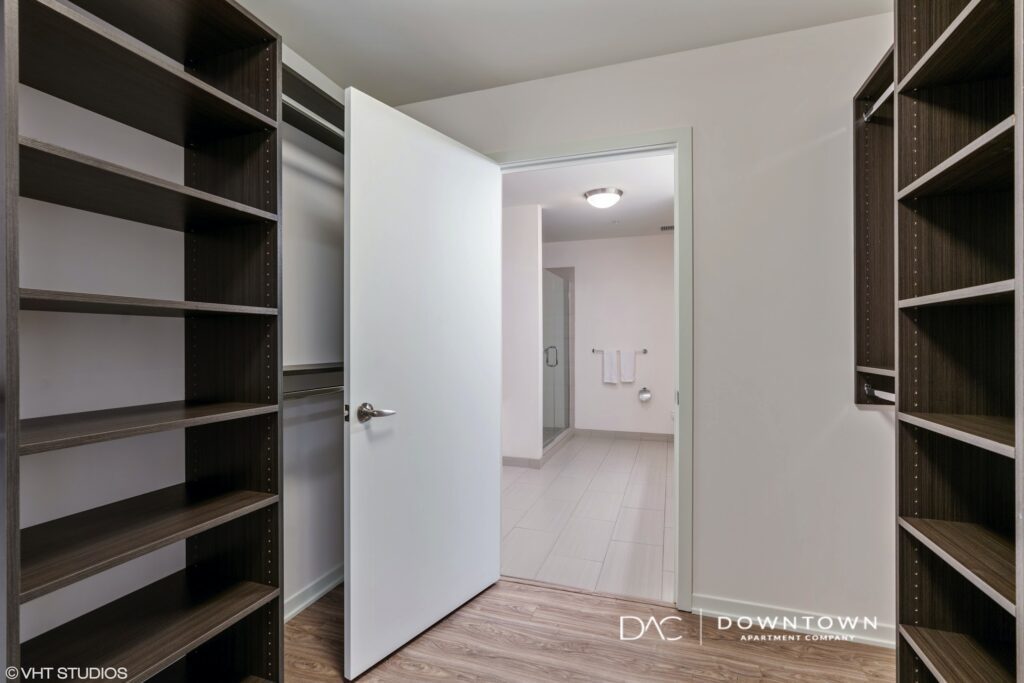 Walk-in closet with empty wooden shelves, an open door leading to a bathroom with tiled floor, and visible towel rail.