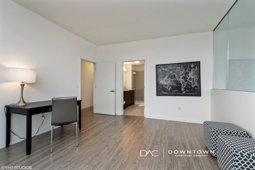 Minimalist room with a desk, chair, and lamp on the left. Open door leading to a bathroom in the center. Black and white map artwork on the wall. Wood flooring and patterned chair on the right.