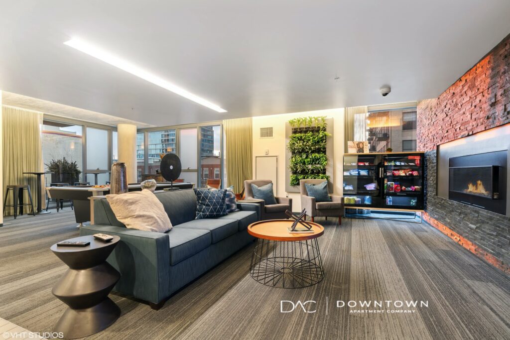 Modern lounge area with sofas, chairs, a round coffee table, wall-mounted TV, and fireplace. Large windows and glass doors provide natural light.
