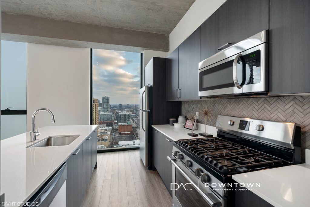 Modern kitchen with dark cabinetry, stainless steel appliances, and a large window with city views. White countertops and herringbone backsplash are visible.
