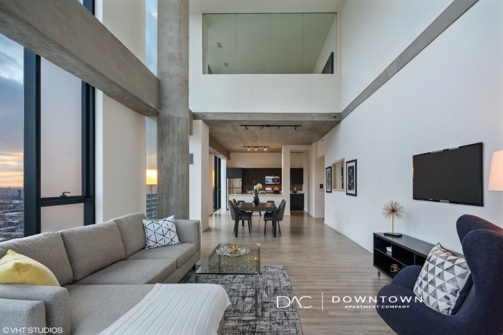 Modern loft apartment interior with high ceilings, large windows, and minimalistic furniture. Living area features a sofa, chairs, and a wall-mounted TV, with a kitchen and dining space in the background.