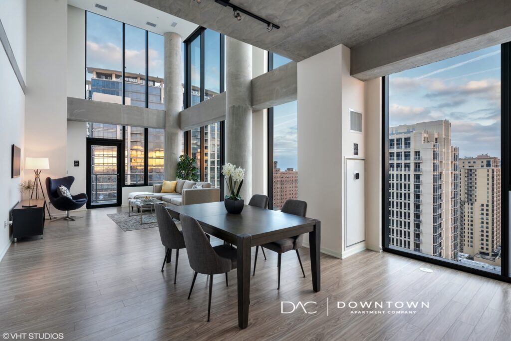 Modern apartment interior with large windows, a dining table, chairs, and a couch. Cityscape visible outside. Light wood flooring and concrete columns enhance the contemporary design.
