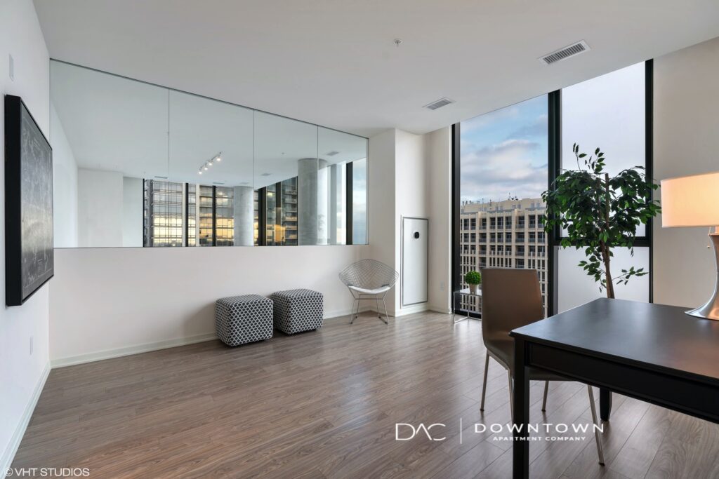Modern office space with hardwood floors, a black desk, lamp, two ottomans, and large mirror reflecting cityscape through floor-to-ceiling windows.