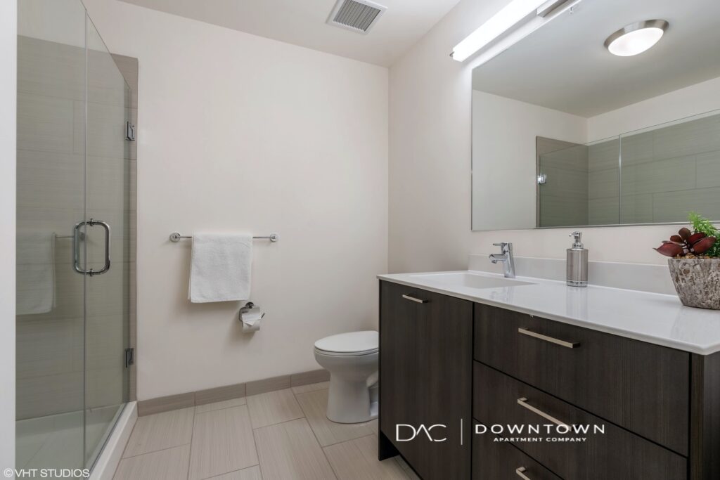 Modern bathroom with a glass-enclosed shower, white toilet, and a dark wood vanity with a white countertop. A towel hangs on a rack, and a potted plant is beside the sink.