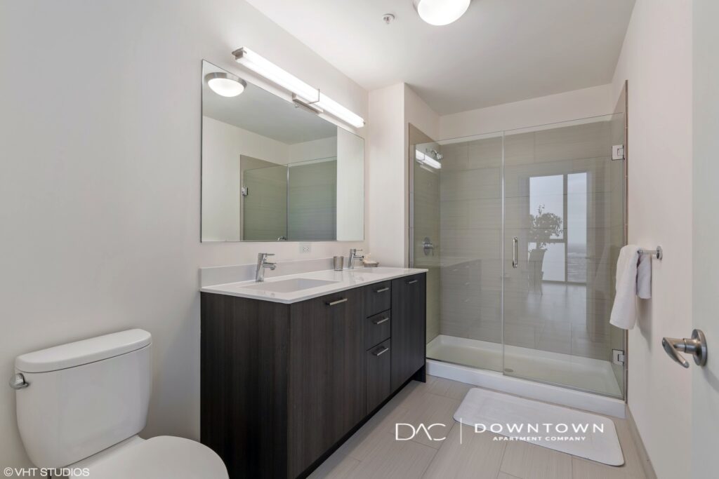 Modern bathroom with a large mirror, double-sink vanity, toilet, and glass-door shower. White walls and sleek fixtures create a clean and minimalistic look.
