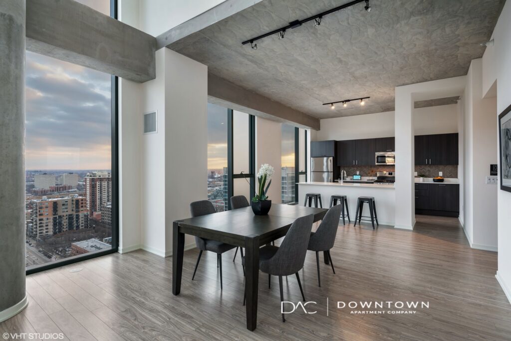 Modern open-plan apartment with tall windows, dining table, and kitchen in view. Cityscape visible through the windows.