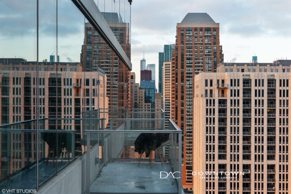 View of a cityscape with tall buildings from a high-rise balcony, featuring a glass railing and two black chairs.
