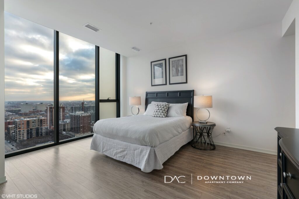 Modern bedroom with large windows, city view, a neatly made bed, two bedside tables with lamps, and framed artwork on the wall. Floor is wooden with a light finish.
