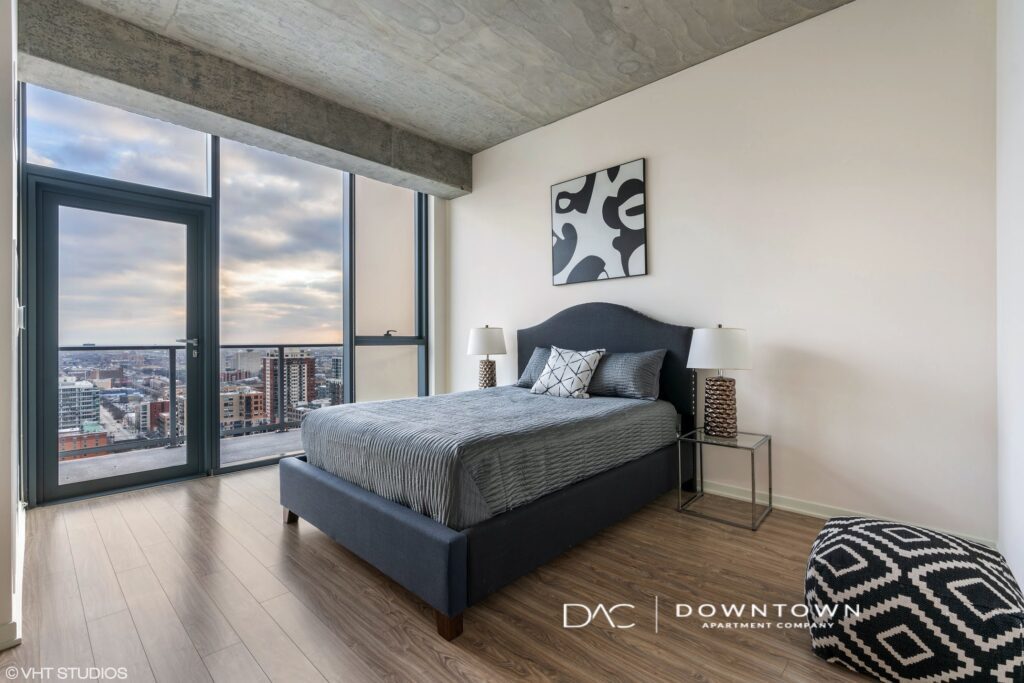 Modern bedroom with a large window view of the city. Features a bed with gray bedding, two lamps, a black and white abstract painting above, and a patterned ottoman.