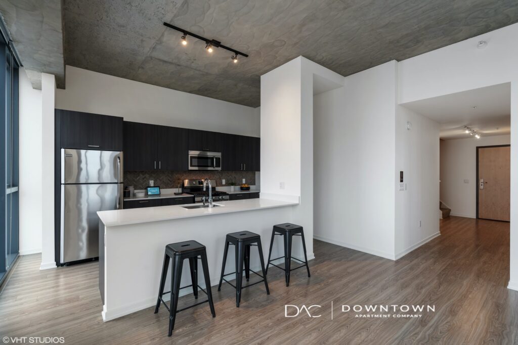 Modern kitchen with stainless steel appliances, dark cabinets, and a large white island with three barstools. Light wood flooring and concrete ceiling.
