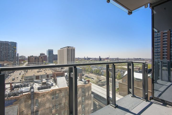Urban landscape view from a balcony with glass railing, overlooking buildings and a clear sky.